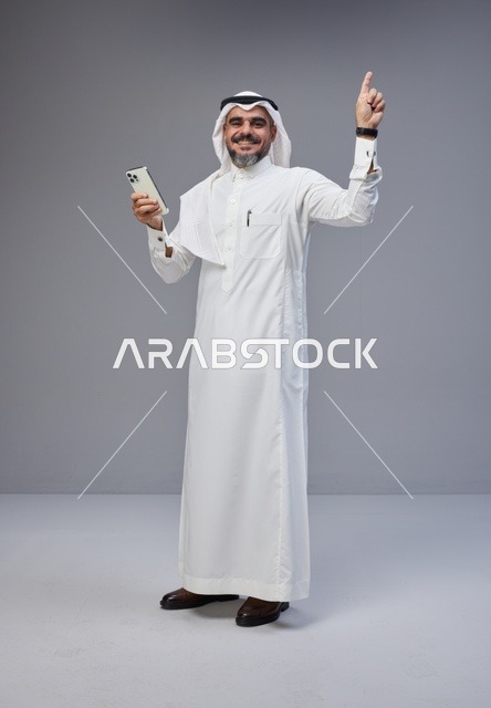 رجل سعودي بالزي التقليدي يشير للأعلى مع هاتف ذكي