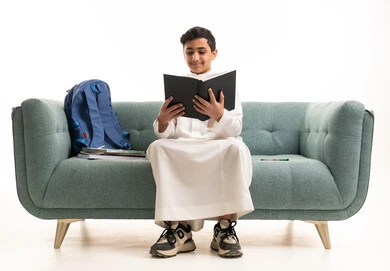 طالب سعودي بالثوب التقليدي يقرأ كتاباً على الأريكة