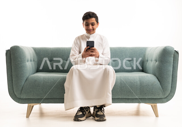طفل سعودي بالثوب التقليدي يستخدم الهاتف الذكي على الأريكة