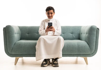 طفل سعودي بالثوب التقليدي يستخدم الهاتف الذكي على الأريكة