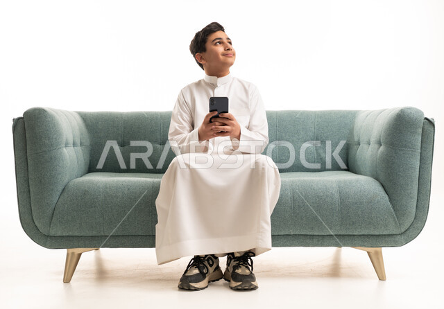 فتى سعودي بالثوب الأبيض يستخدم الهاتف الذكي على الأريكة