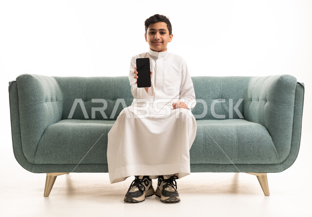 فتى سعودي بالثوب يحمل هاتفاً ذكياً على أريكة بخلفية بيضاء