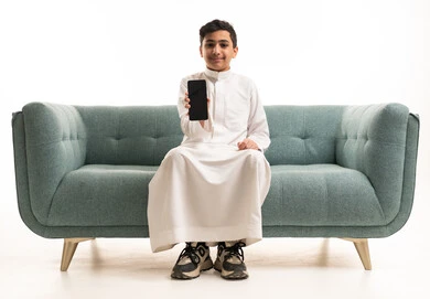 فتى سعودي بالثوب يحمل هاتفاً ذكياً على أريكة بخلفية بيضاء