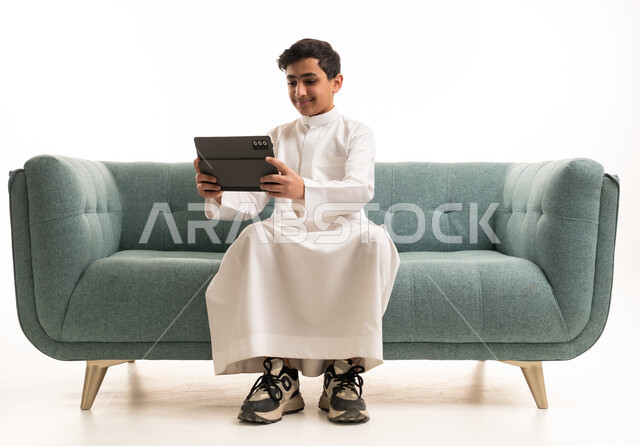 فتى سعودي بالثوب يستخدم جهازاً لوحياً على الأريكة