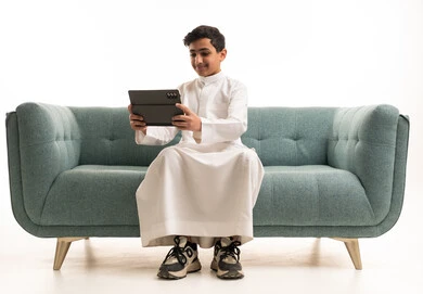 فتى سعودي بالثوب يستخدم جهازاً لوحياً على الأريكة