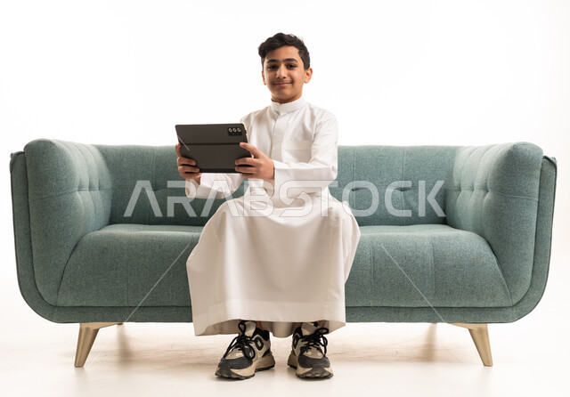 فتى سعودي بالثوب يمسك جهاز لوحي على الأريكة