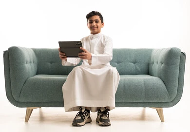 فتى سعودي بالثوب يمسك جهاز لوحي على الأريكة