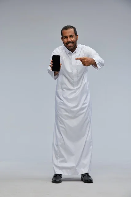 رجل سعودي بالثوب الأبيض يشير إلى شاشة هاتف ذكي
