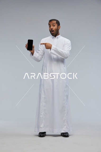 رجل سعودي بالثوب الأبيض يشير إلى شاشة هاتف ذكي
