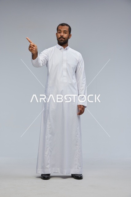 رجل سعودي يرتدي الثوب الأبيض ويشير أمام خلفية رمادية