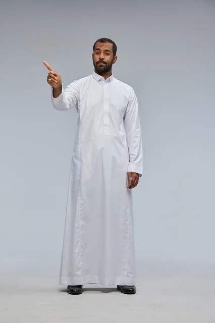 رجل سعودي يرتدي الثوب الأبيض ويشير أمام خلفية رمادية