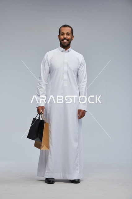 رجل سعودي يرتدي الثوب الأبيض ويحمل أكياس تسوق ملونة