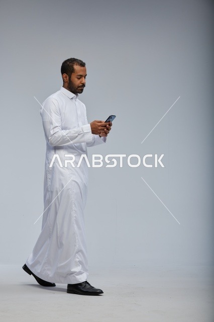 Saudi Man in White Thobe Using Smartphone Walking