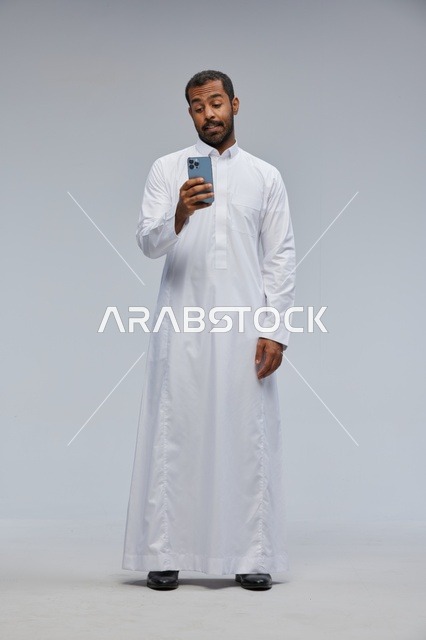 رجل سعودي بالثوب الأبيض يستخدم هاتف ذكي في الاستوديو