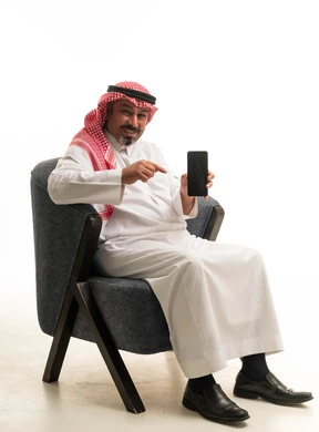 رجل سعودي في الزي التقليدي يشير إلى هاتف ذكي بخلفية بيضاء