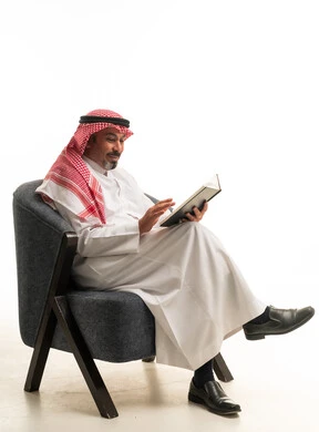 رجل سعودي بالزي التقليدي يقرأ كتاباً على كرسي مريح