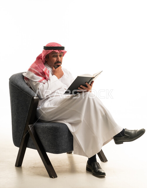 رجل سعودي بالثوب يقرأ كتاباً في الاستوديو على خلفية بيضاء