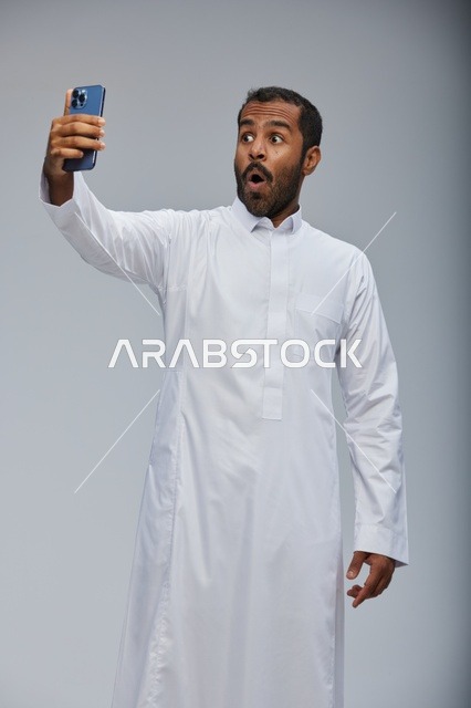رجل سعودي بالثوب الأبيض يمسك هاتف بتعبير مندهش