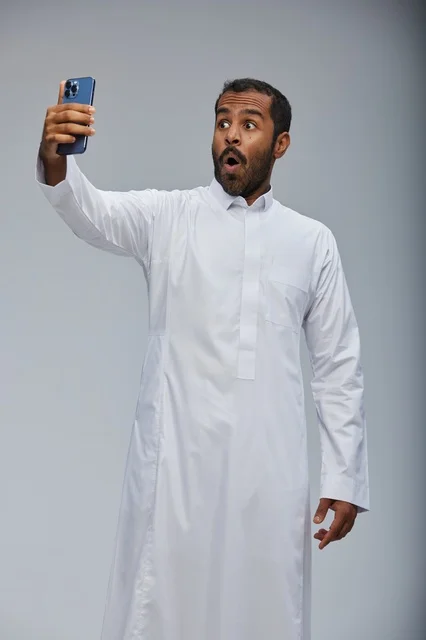 رجل سعودي بالثوب الأبيض يمسك هاتف بتعبير مندهش