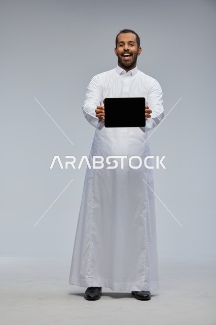 رجل سعودي بالثوب الأبيض يمسك جهازاً لوحياً في الاستوديو
