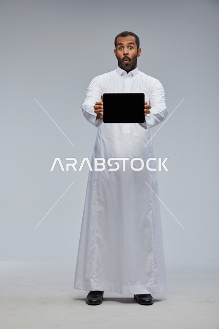 رجل سعودي بالثوب الأبيض يحمل جهاز لوحي رقمي بتعبير متفاجئ