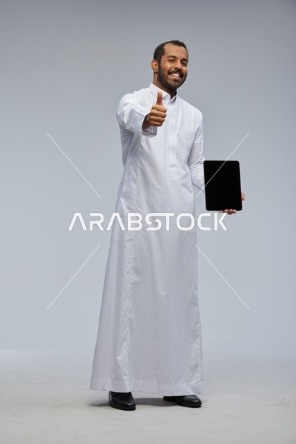 رجل سعودي بالثوب الأبيض يحمل جهاز لوحي وإبهام للأعلى