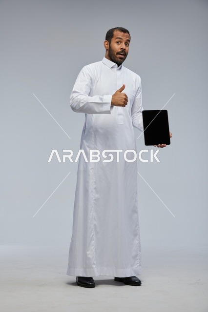 رجل سعودي بالثوب الأبيض يحمل جهاز لوحي مع إيماءة إعجاب