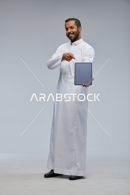 رجل سعودي بالثوب الأبيض يعرض جهازاً لوحياً في الاستوديو