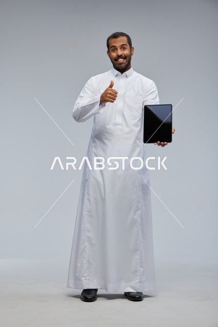رجل سعودي بالثوب الأبيض يحمل جهازاً لوحياً وإيماءة نجاح