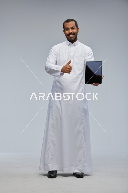 رجل سعودي بالثوب الأبيض يمسك جهاز لوحي مبتسماً