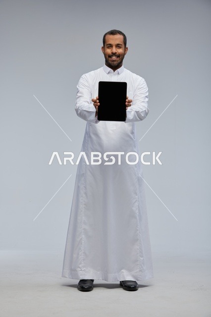 رجل سعودي بالثوب الأبيض يمسك جهازاً لوحياً في الاستوديو