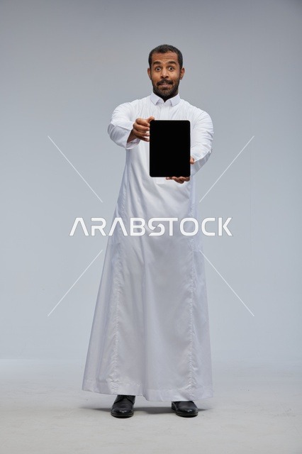 رجل سعودي بالثوب الأبيض يحمل جهاز لوحي في استوديو