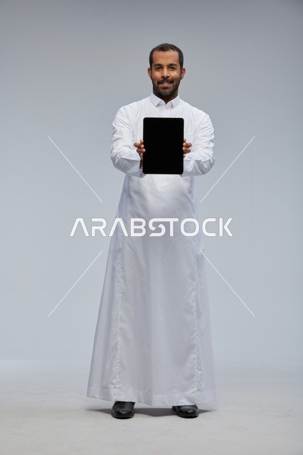 رجل سعودي بالثوب الأبيض يمسك جهازاً لوحياً في الاستوديو