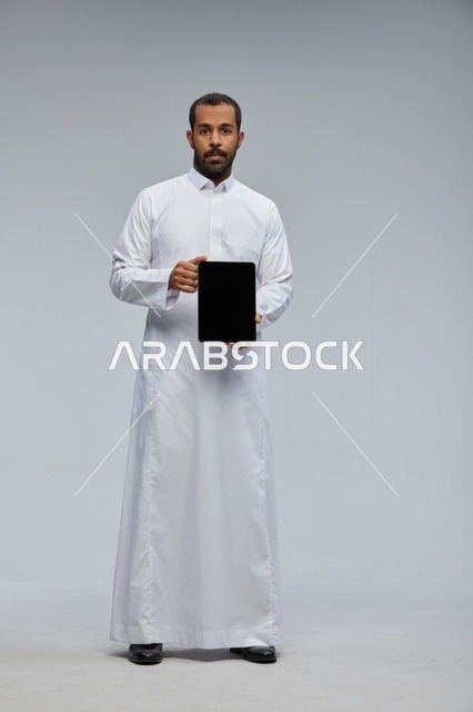 رجل سعودي بالثوب الأبيض يحمل جهازاً لوحياً في الاستوديو