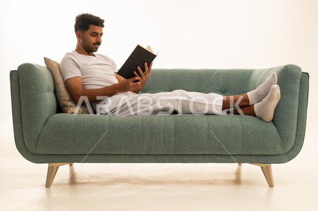 رجل سعودي يقرأ كتاباً على أريكة خضراء بخلفية بيضاء