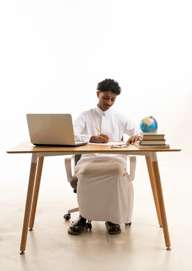 Saudi Male Student Writing at Desk with Laptop