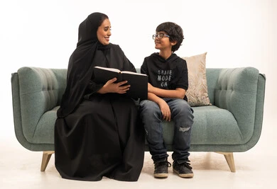 امرأة سعودية بالعباءة تقرأ كتاباً مع ابنها على الأريكة