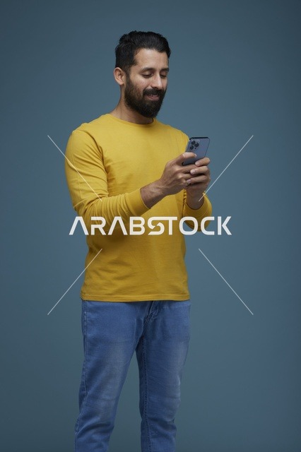 شاب سعودي بملابس كاجوال يستخدم هاتف ذكي أمام خلفية زرقاء