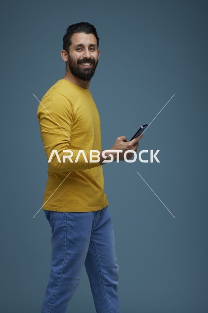 شاب سعودي يبتسم يحمل هاتف ذكي في استوديو بخلفية زرقاء