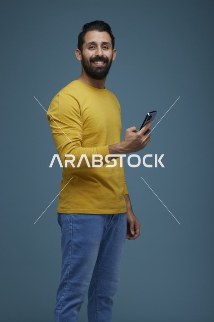 شاب سعودي مبتسم يحمل هاتفاً ذكياً على خلفية زرقاء