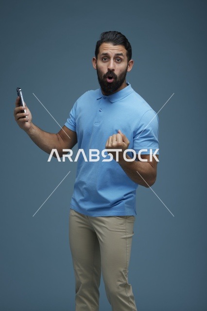 شاب سعودي مندهش يحمل هاتفا ذكيا خلفية زرقاء