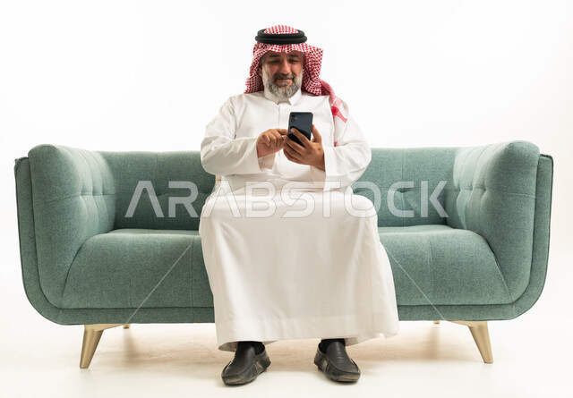 رجل سعودي بالثوب التقليدي يستخدم الهاتف على الأريكة