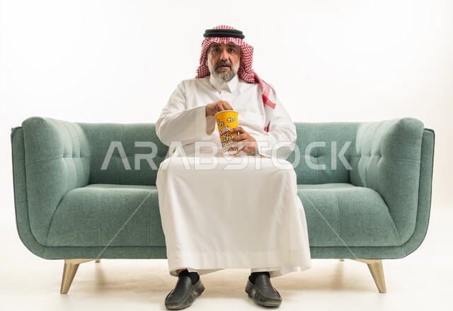 رجل سعودي بالثوب التقليدي يأكل الفشار على الأريكة