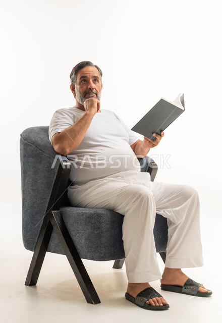 رجل سعودي يقرأ كتاباً على كرسي مريح بخلفية بيضاء