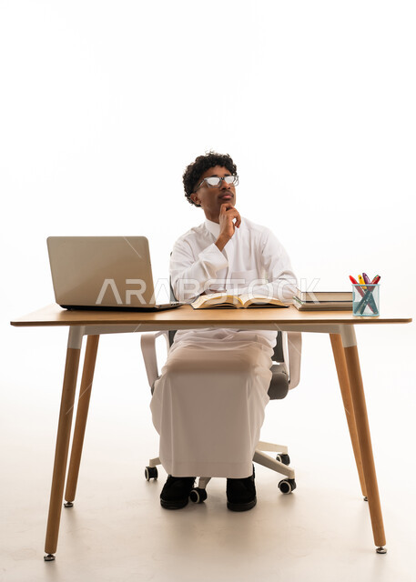 رجل سعودي يرتدي الثوب التقليدي يفكر خلف مكتب العمل بالاستوديو