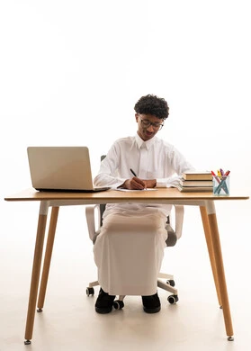Young Saudi Student Writing at Desk with Laptop