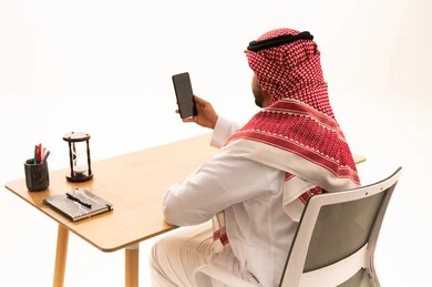 رجل سعودي بالثوب والشماغ في مكتبه يستخدم الهاتف الذكي