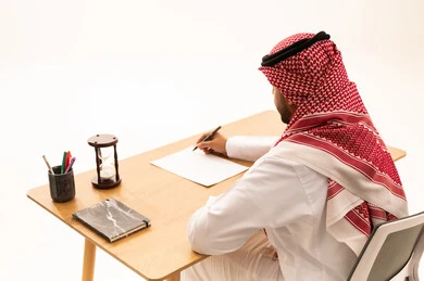 رجل سعودي يكتب على مكتب خشبي مع ساعة رملية