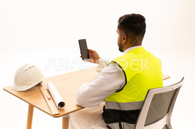 مهندس سعودي يجلس خلف مكتبه مع هاتف ذكي وخوذة سلامة