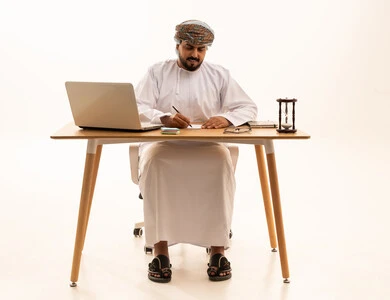 Omani Businessman Writing at Desk with Laptop